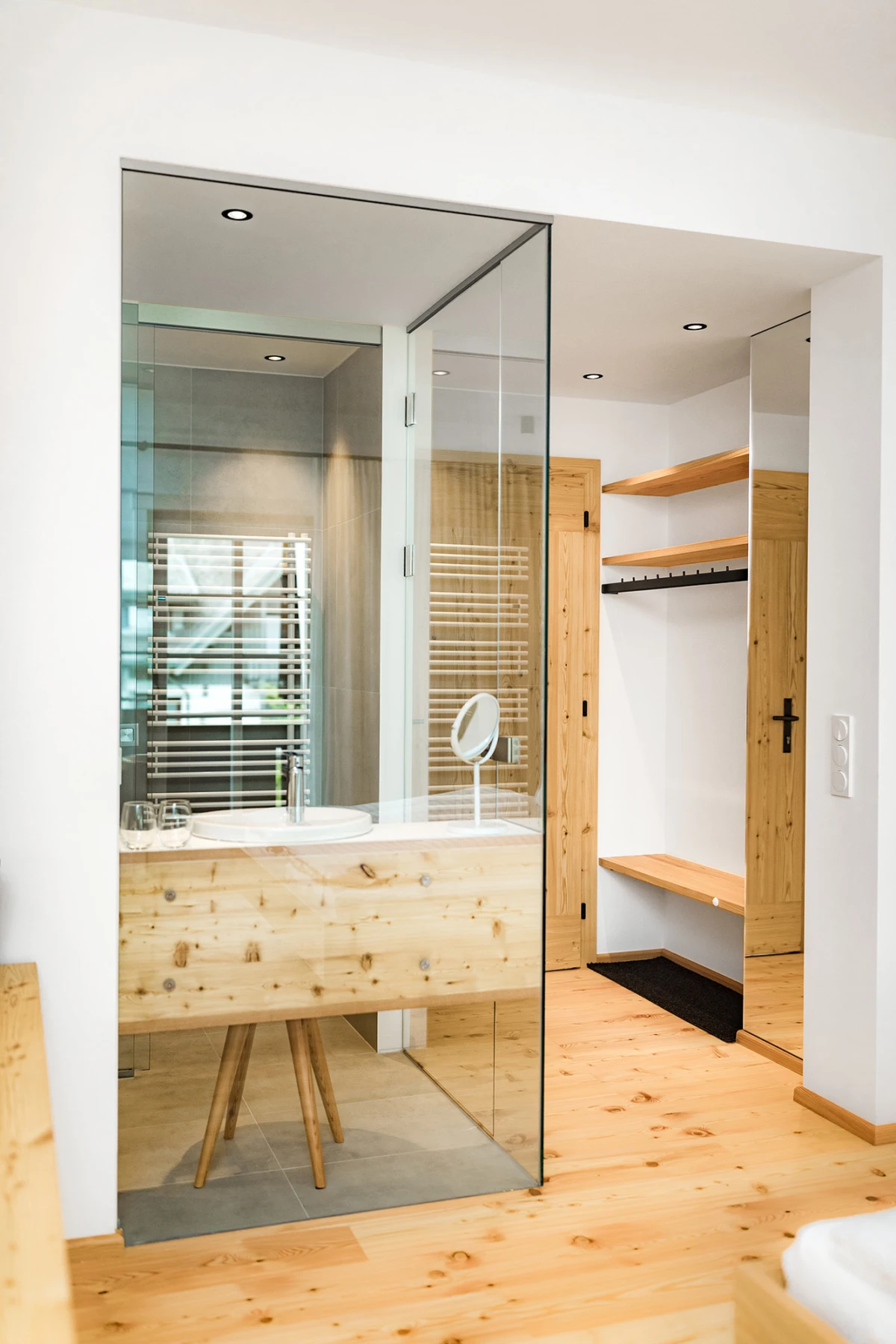 Offenes Badezimmer hinter Glaswand: Holz-Waschtisch mit rundem Spiegel, Holzfußboden; angrenzende Garderobe mit Regal und Haken.
