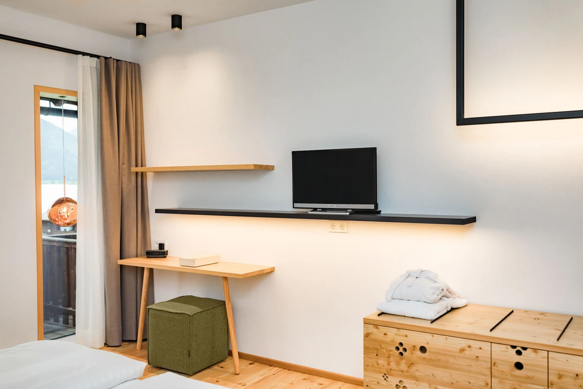 Modernes Hotelzimmer: Wand-TV auf schwarzem Regal, Holzschreibtisch, grüner Pouf, Holzkommode mit Handtüchern, Balkon mit Bergblick.
