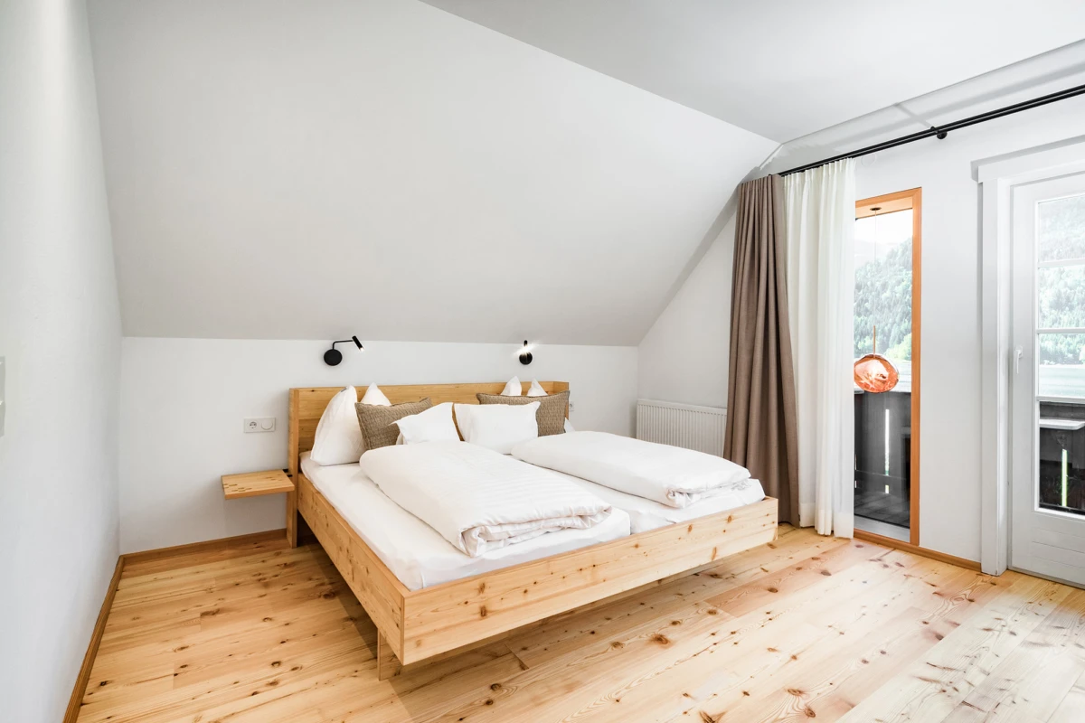 Helles Schlafzimmer mit hellem Holzbett, weißer Bettwäsche und Kissen; zwei schwarze Wandlampen, Holzboden, Balkonzugang mit Vorhängen.