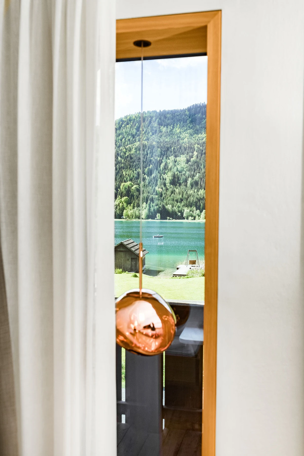 Kupferne Pendellampe hängt vor Fensterrahmen; Blick auf See, Bootshaus und Wald im Hintergrund.