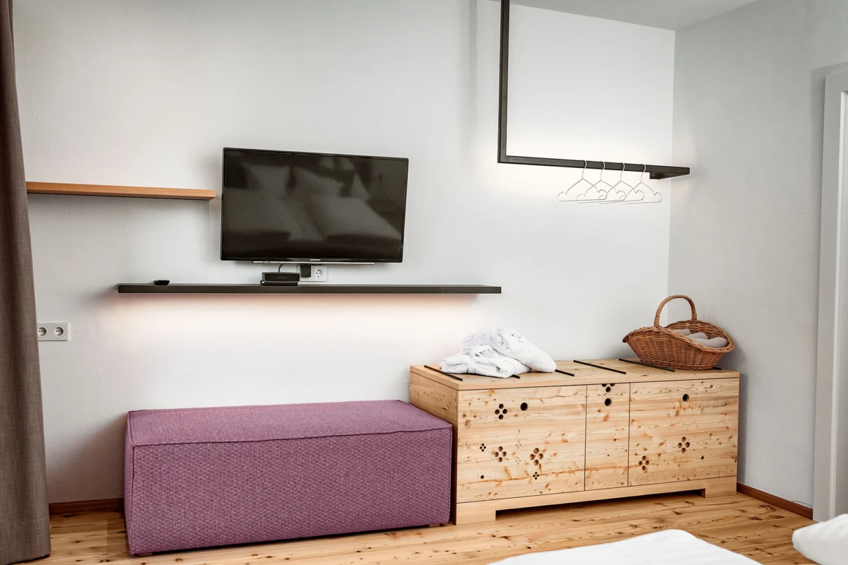 Hotelzimmer mit Wandfernseher über schmaler Ablage, violetter Pouf, Holzschrank mit Handtüchern und Korb auf dem Schrank, Holzboden.