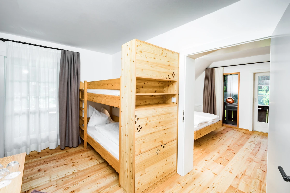 Kiefernholz-Etagenbett in hellem Gästezimmer mit Holzboden; offene Tür führt zu weiterem Schlafzimmer mit Einzelbett.