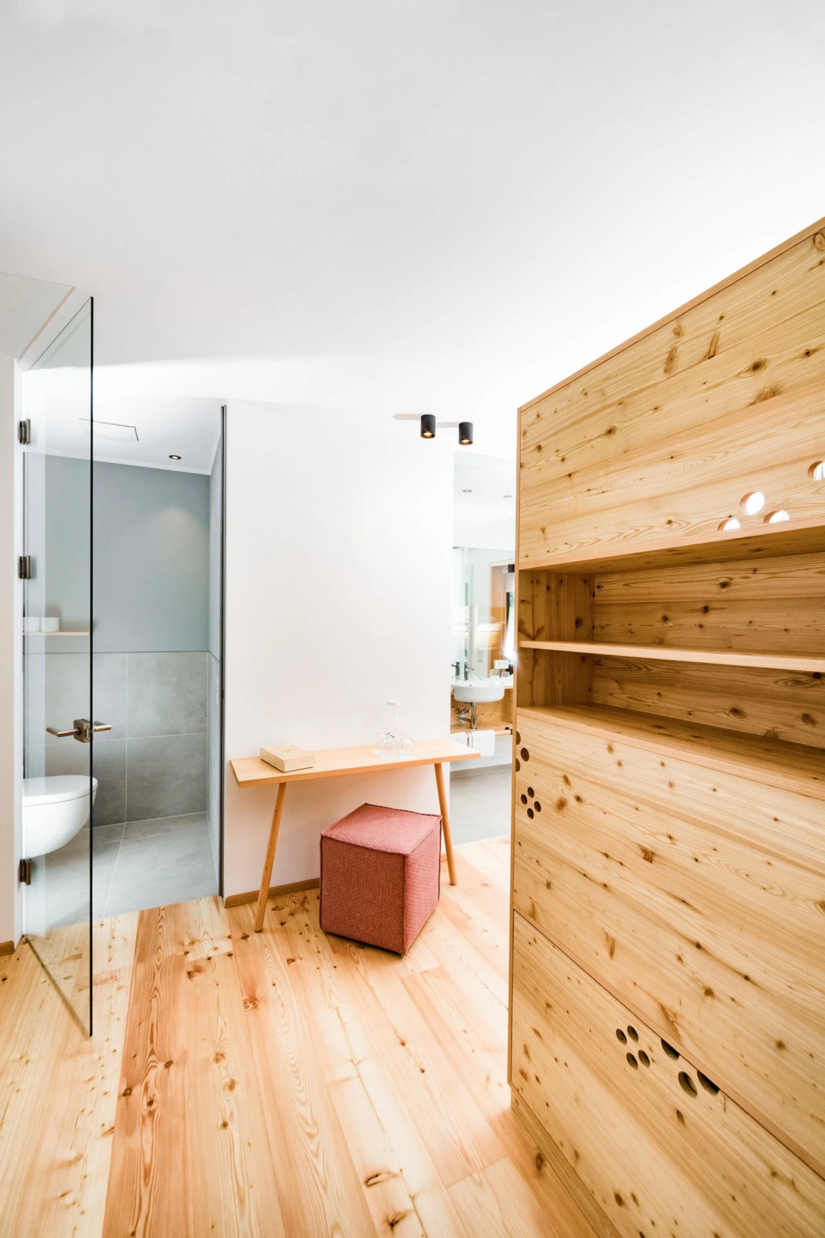 Moderne Innenraumszene: Holzfußboden, großer Holzschrank rechts, Glastür zum Badezimmer links mit Toilette, schmaler Holztisch und roter Würfelhocker.