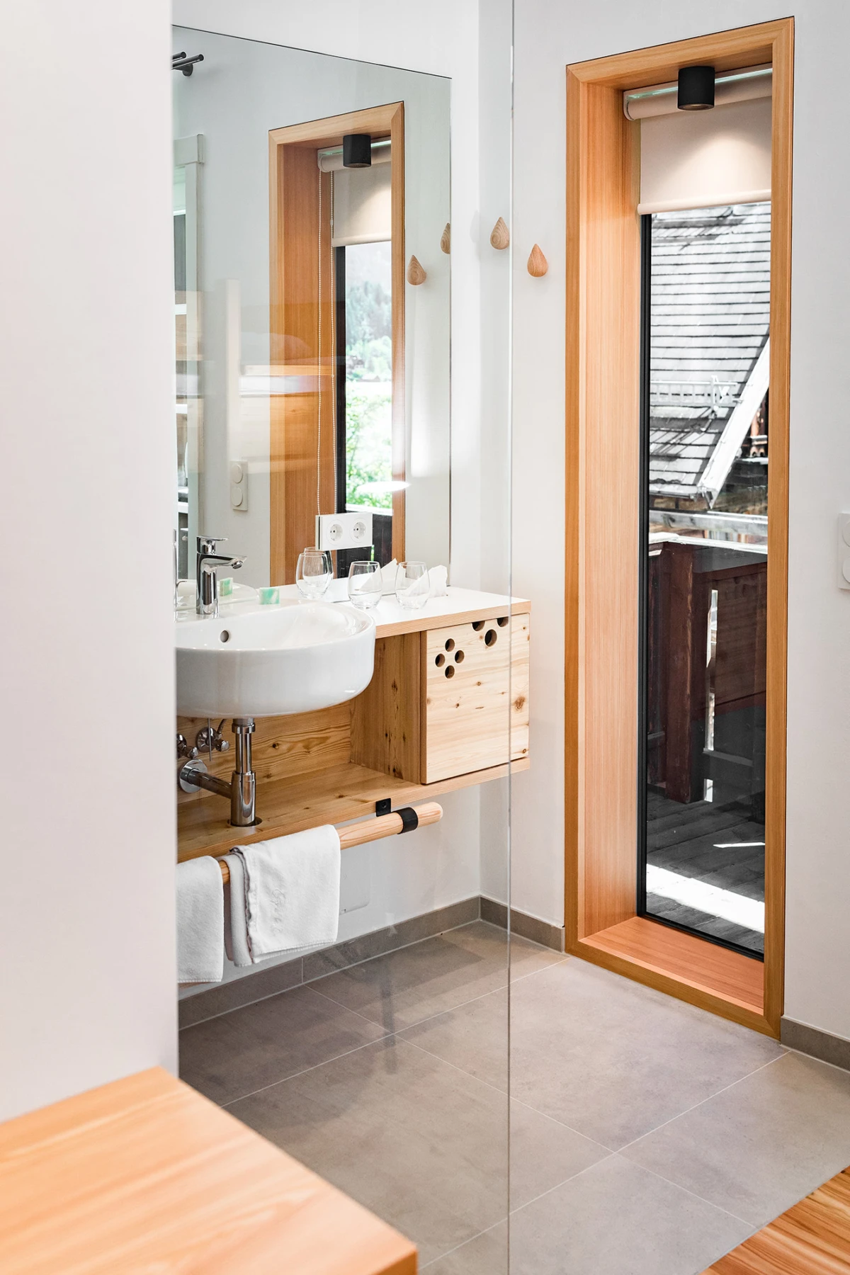 Modernes Badezimmer: Waschbecken auf Holzablage, Holzkasten, Glaswand, Handtuchstange; Fensterfront nach draußen sichtbar.