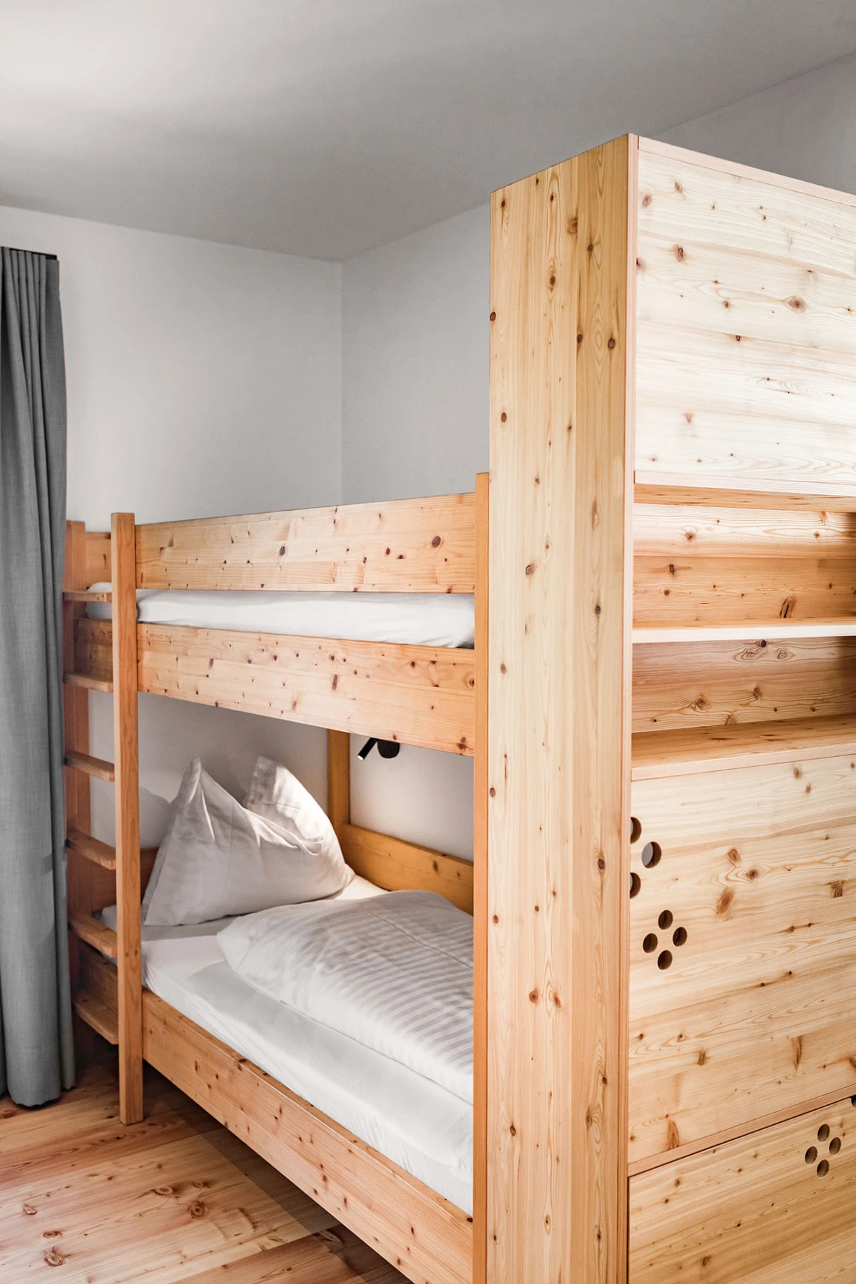Holz-Etagenbett im Schlafzimmer; unbesetzte obere und untere Liegeflächen mit weißen Bettlaken, grauer Vorhang links.