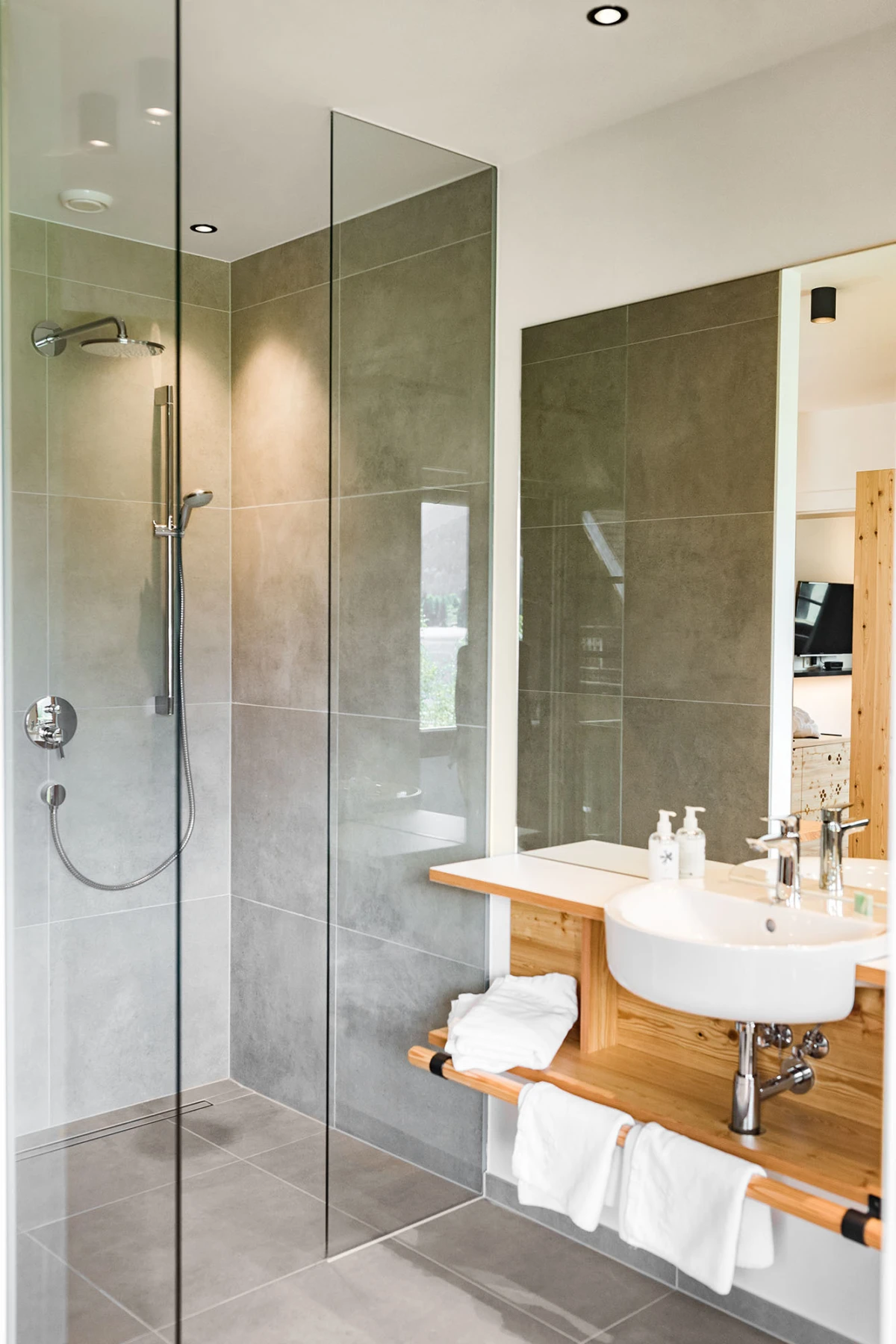 Modernes Badezimmer mit Glasdusche, grauen Fliesen, großem Spiegel und Waschbecken auf Holzablage, Handtücher darunter.