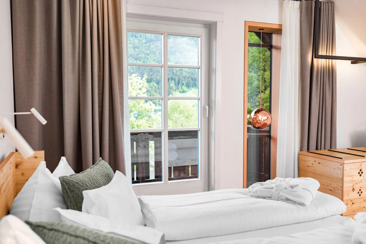 Modernes Schlafzimmer: Bett mit weißer Bettwäsche und grauen Kissen, Balkonfenster mit Blick auf grüne Landschaft, kupferne Pendelleuchte.