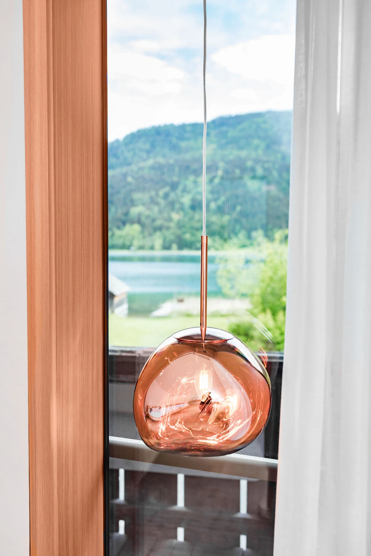 Roségoldene Glas-Pendelleuchte hängt vor einem Fenster; draußen Blick auf See und grüne Berge, Holzrahmen, weiße Vorhänge.