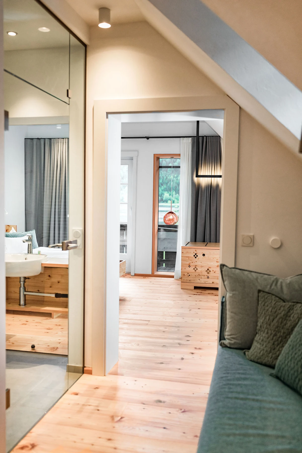 Heller Innenraum: Flur zum Schlafzimmer mit Holzfußboden, grauen Vorhängen, roter Pendelleuchte und Pfotenmuster-Holzkasten; Sofa rechts.