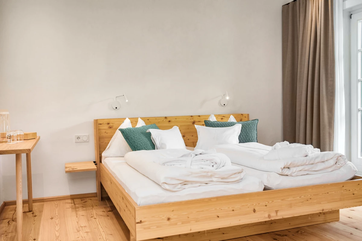 Massivholz-Bett mit weißer Bettwäsche und grünen Kissen in hellem Schlafzimmer; beige Vorhänge, Fenster rechts.