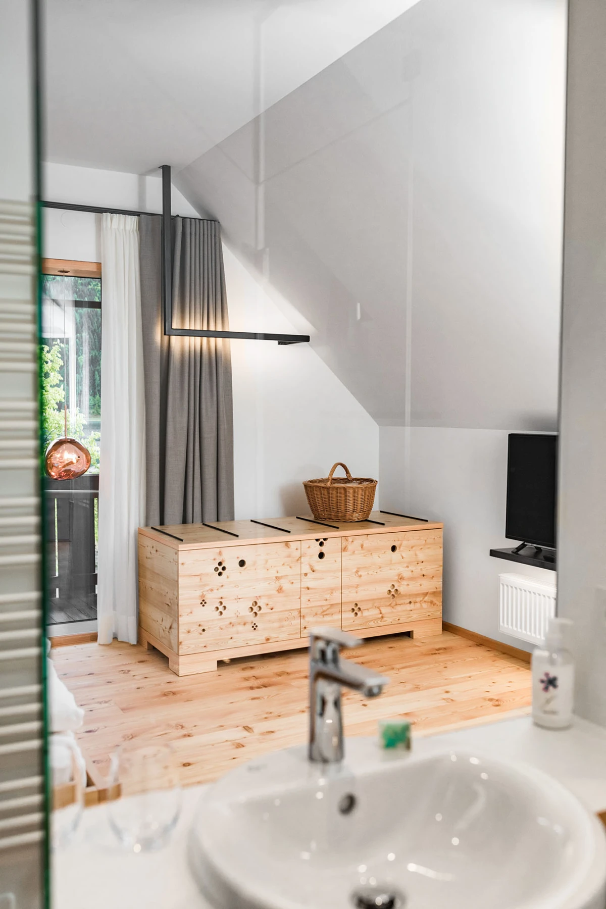 Moderne Innenansicht: Holzkommode mit Pfotenspuren und Korb oben, TV rechts, Balkonfenster mit Vorhängen; Waschbecken im Vordergrund.