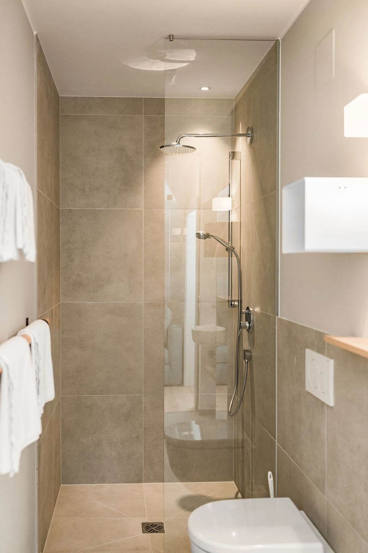 Modernes Badezimmer mit Glasdusche, Regen- und Handbrause, beigen Fliesen, Toilette rechts, Handtücher links.