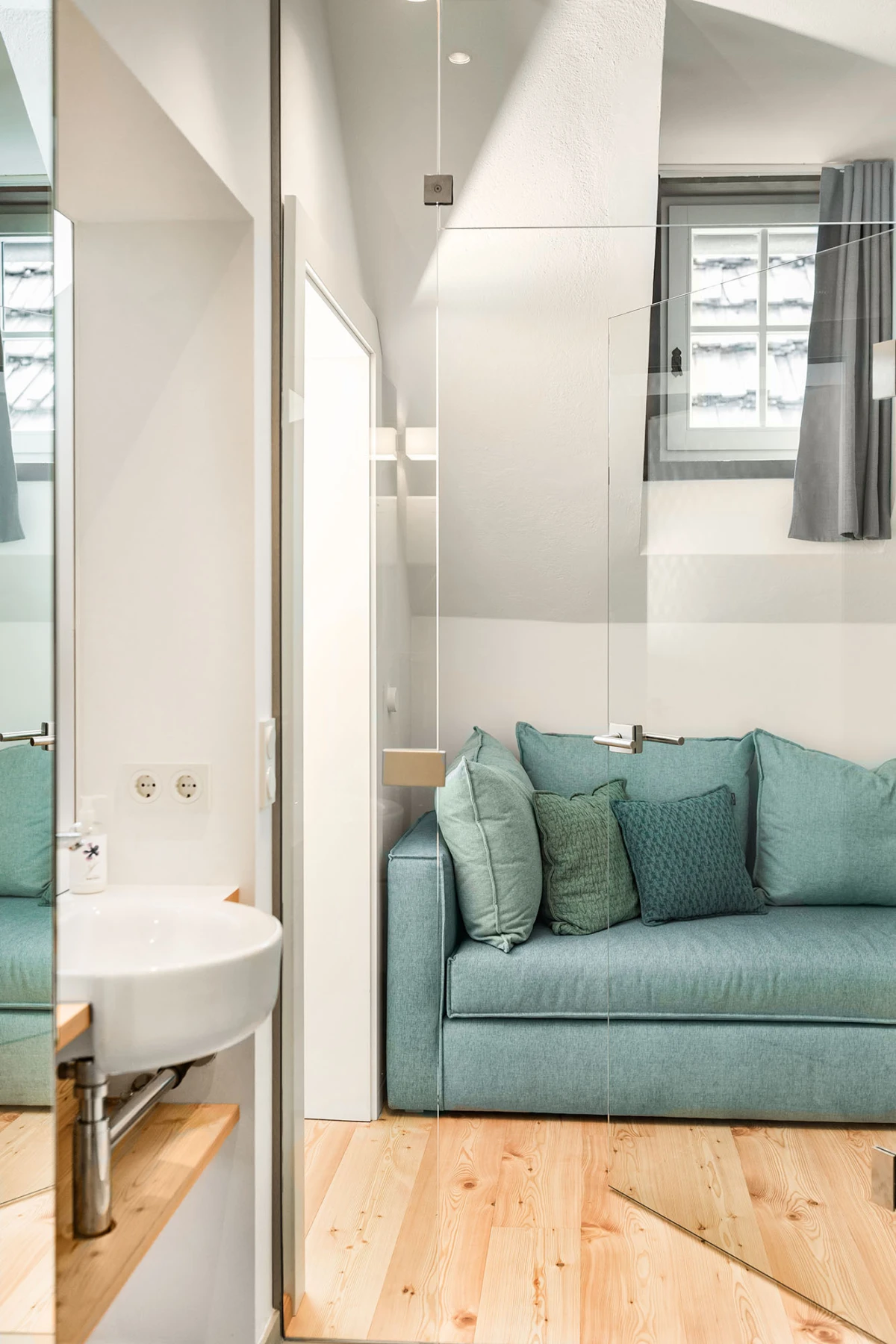 Türkisfarbene Couch mit Kissen im hellen Wohnzimmer; Glaswand, Holzboden, Fenster mit Vorhängen.