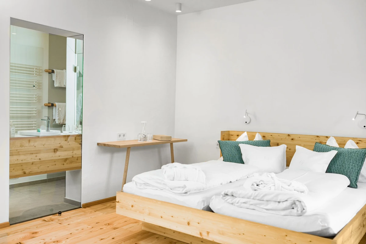 Schlafzimmer mit Holzplattformbett, weißer Bettwäsche und grünen Kissen; Holztisch daneben; offenes Badezimmer mit Holzmöbeln und Handtüchern.