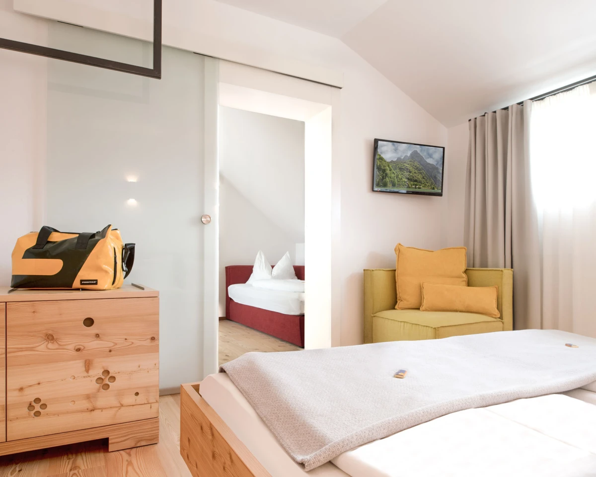 Hotelzimmer mit Bett im Vordergrund, gelber Reisetasche auf Holzmöbeln, gelber Sessel, Wandfernseher mit Berglandschaft, Fenster mit Vorhang.