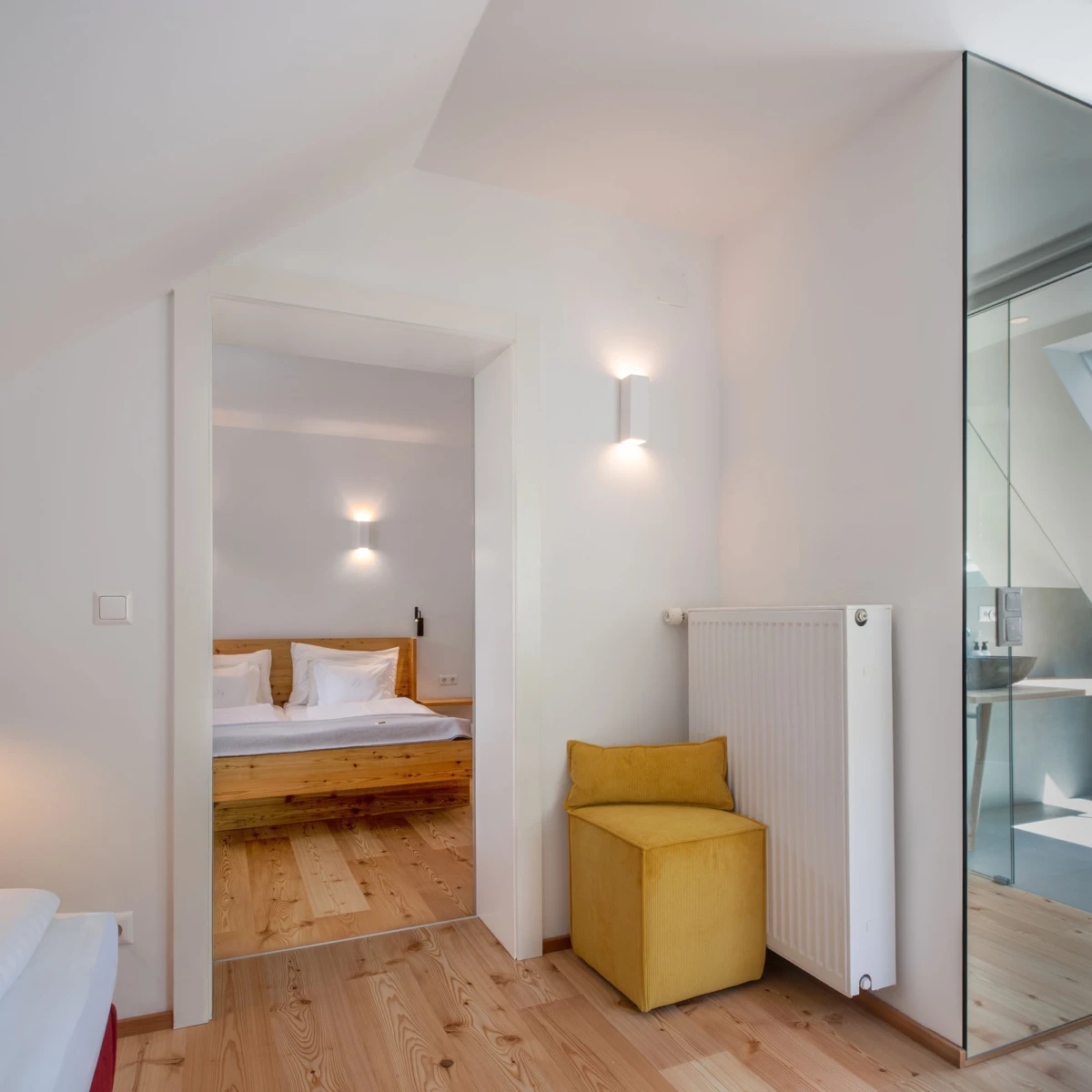 Modernes Schlafzimmer mit Holzbett und weißer Bettwäsche, gelbem Pouf neben Heizkörper; offener Durchgang zu zweitem Schlafzimmer.