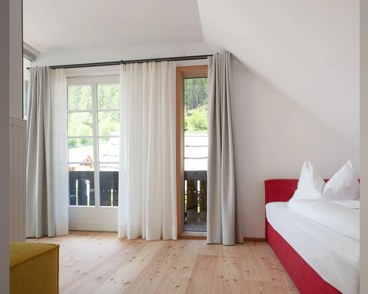 Schlafzimmer mit rotem Doppelbett, weißer Bettwäsche, hellen Vorhängen, Holzboden; Blick auf Balkon und grüne Landschaft.