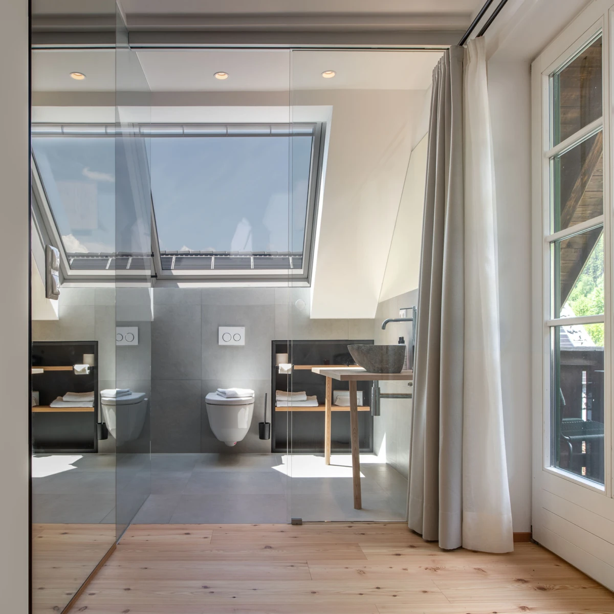 Modernes Loft-Badezimmer mit Glasabtrennung, wandhängende Toilette, Steinwaschbecken auf Holzständer, Holzboden, Dachfenster und Vorhänge.