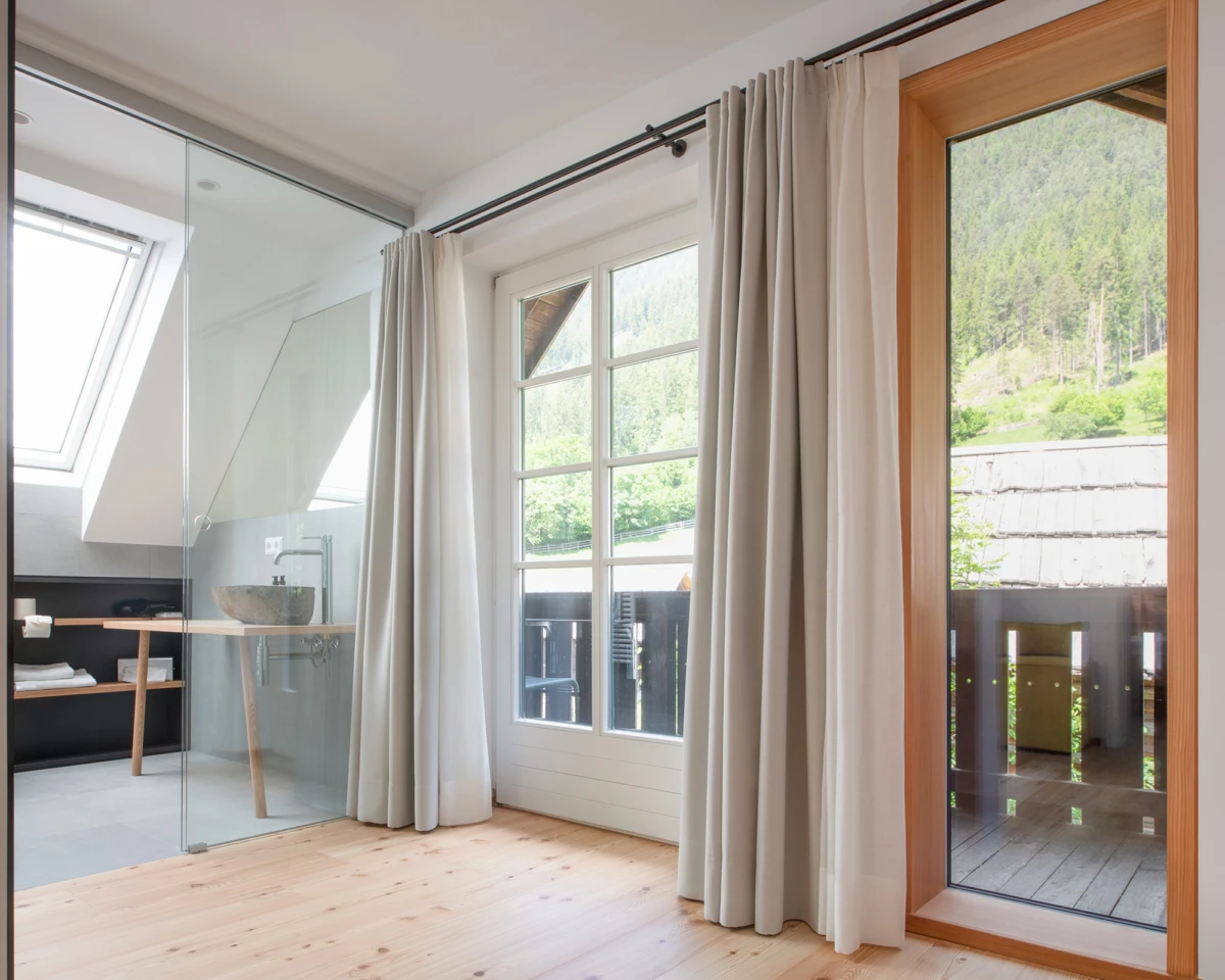 Moderner Innenraum mit Glastrennwand, Steinwaschbecken auf Holzkonsole, große Fenster und Vorhänge, Blick nach Balkon ins Grünland.