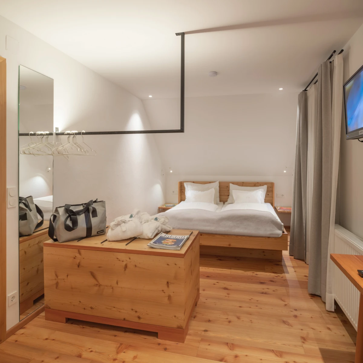 Doppelbett in hellem Hotelzimmer mit Holzmöbeln, Spiegel, Gepäck auf Truhe, Gardinen, Wandfernseher.
