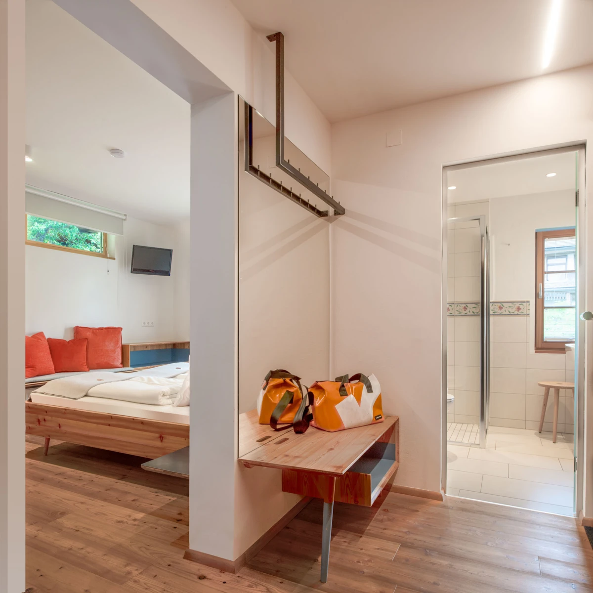 Flurbereich zwischen Schlafzimmer und Badezimmer; Holzbank mit orangefarbenen Taschen, Bett mit roten Kissen im Hintergrund.