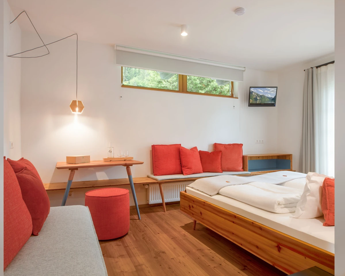 Schlafzimmer mit Holzbett, weißer Bettwäsche und roten Kissen; Sitzbank mit roten Kissen, Wand-TV, Fensterfront mit Vorhängen, Holzfußboden