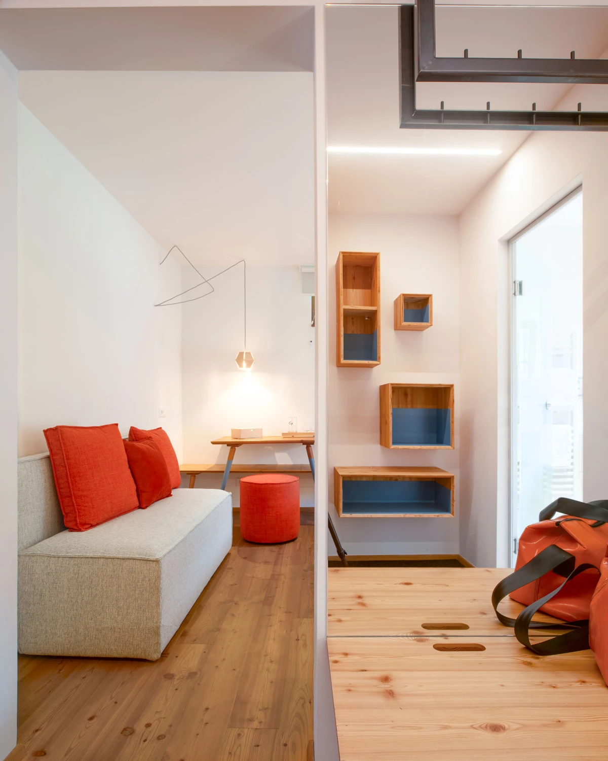 Wohnzimmer-Ecke: graues Ecksofa mit roten Kissen, Schreibtisch mit Lampe, roter Pouf, hölzerne Regale, große Fensterfront.