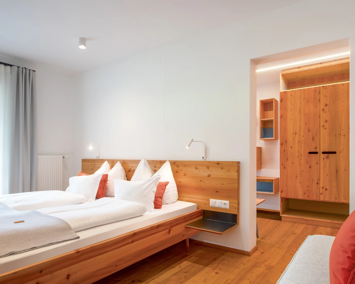 Holz-Doppelbett mit weißen und roten Kissen in hellem Hotelzimmer; Holzfußboden und offener Kleiderschrank im Hintergrund.