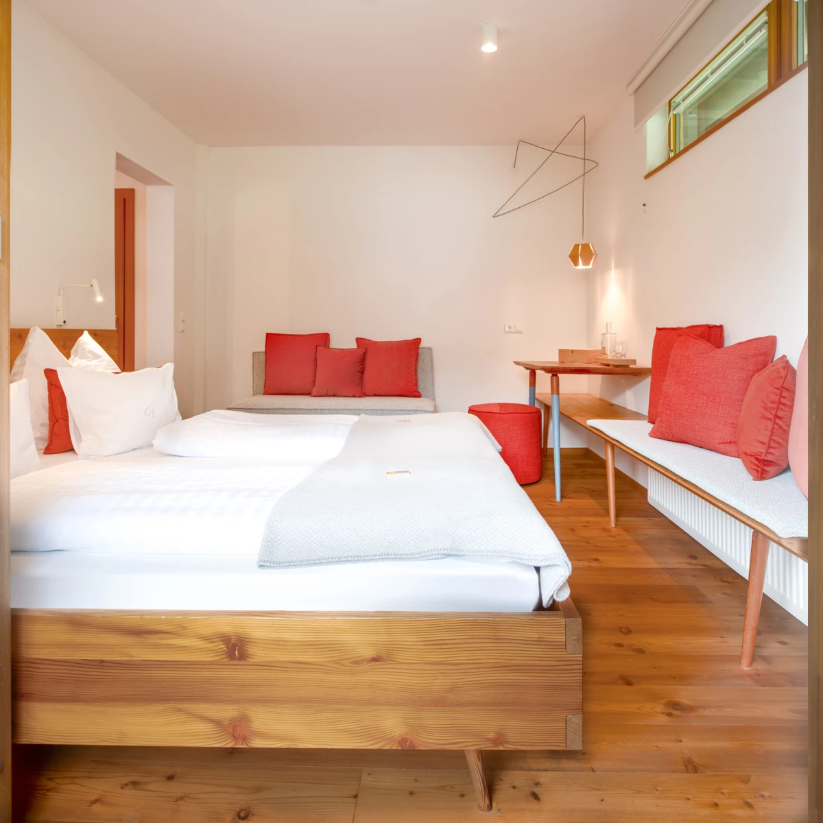 Großes Holzbett im Hotelzimmer mit weißer Bettwäsche und roten Kissen; Sitzecke, Schreibtischbereich, Holzfußboden.