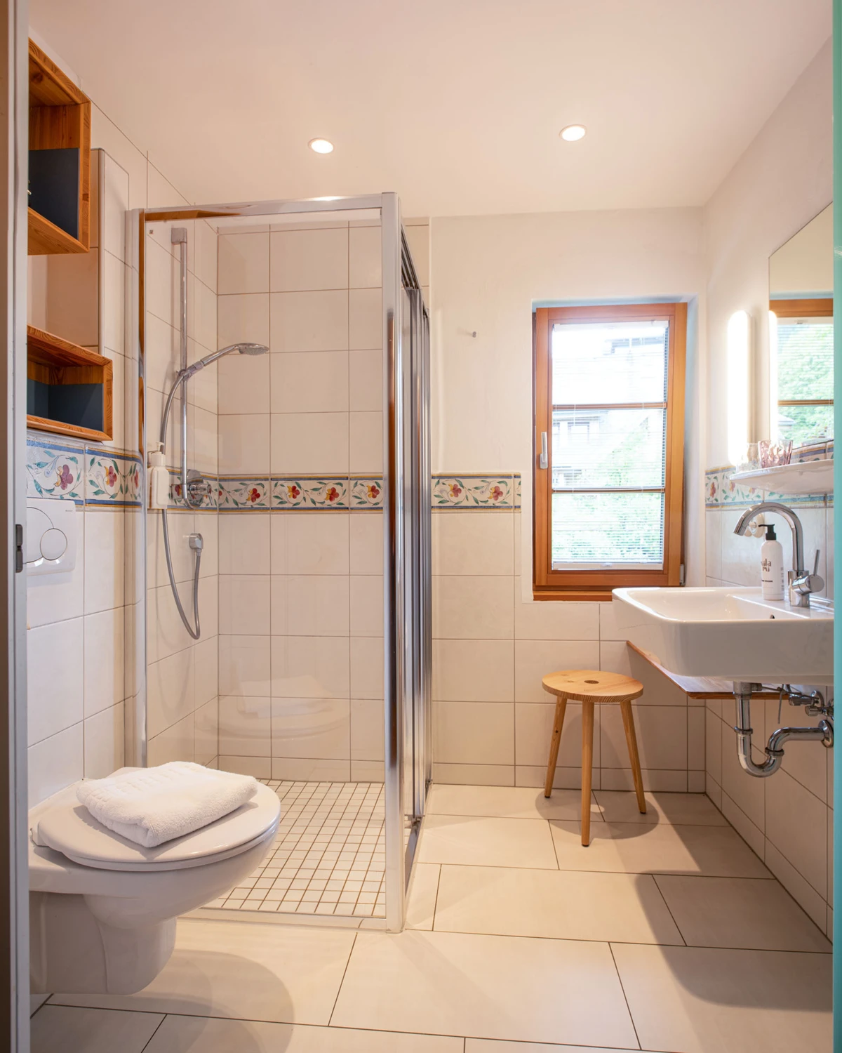 Helles Badezimmer mit Glas-Duschkabine, Toilette mit Handtuch, Waschbecken, Holzfenster und kleinem Holzhocker; florale Bordüre an Wandfliesen.