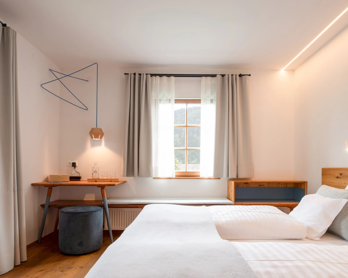 Schlafzimmer mit Bett rechts, Schreibtisch links, Fenster mit Vorhängen, runder Hocker, warmer Holzboden, dezente Beleuchtung.