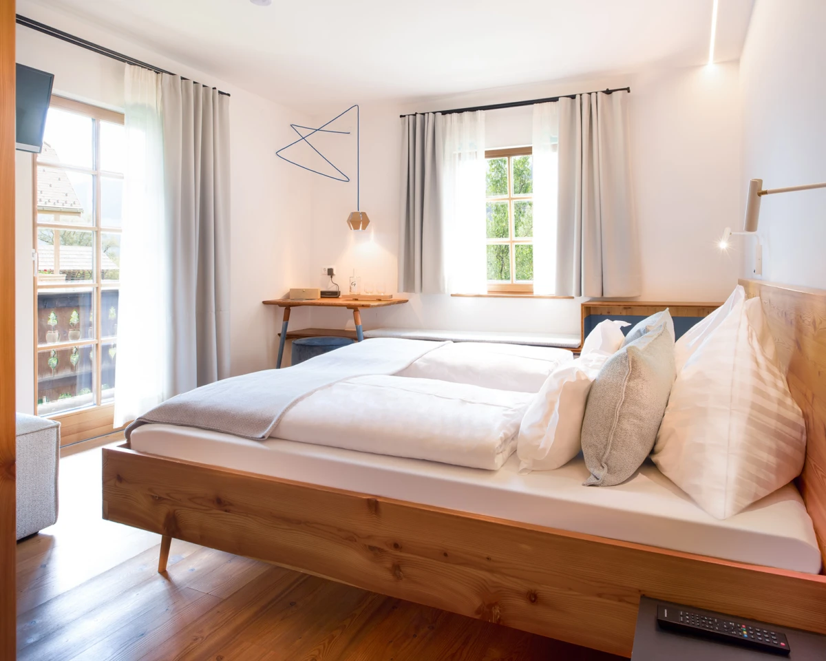 Helles Schlafzimmer mit massivem Holzbett, weißer Bettwäsche und grauen Kissen; Schreibtisch am Fenster, Vorhänge, warme Beleuchtung.