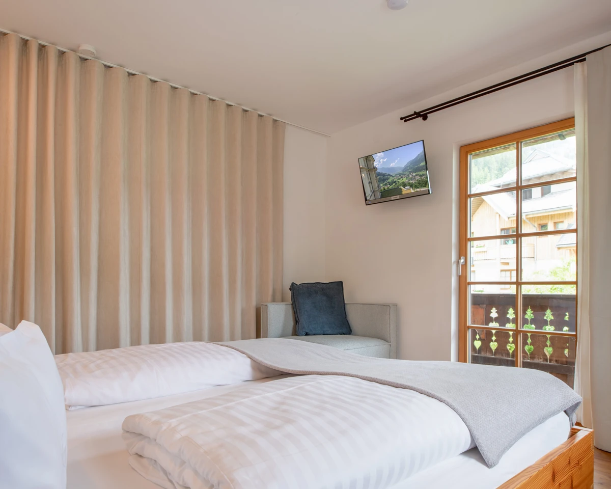 Schlafzimmer mit Doppelbett, weißer Bettwäsche, grauer Überwurf; beige Vorhänge, Wandfernseher, Sofa mit blauem Kissen; Balkontür zum Balkon.
