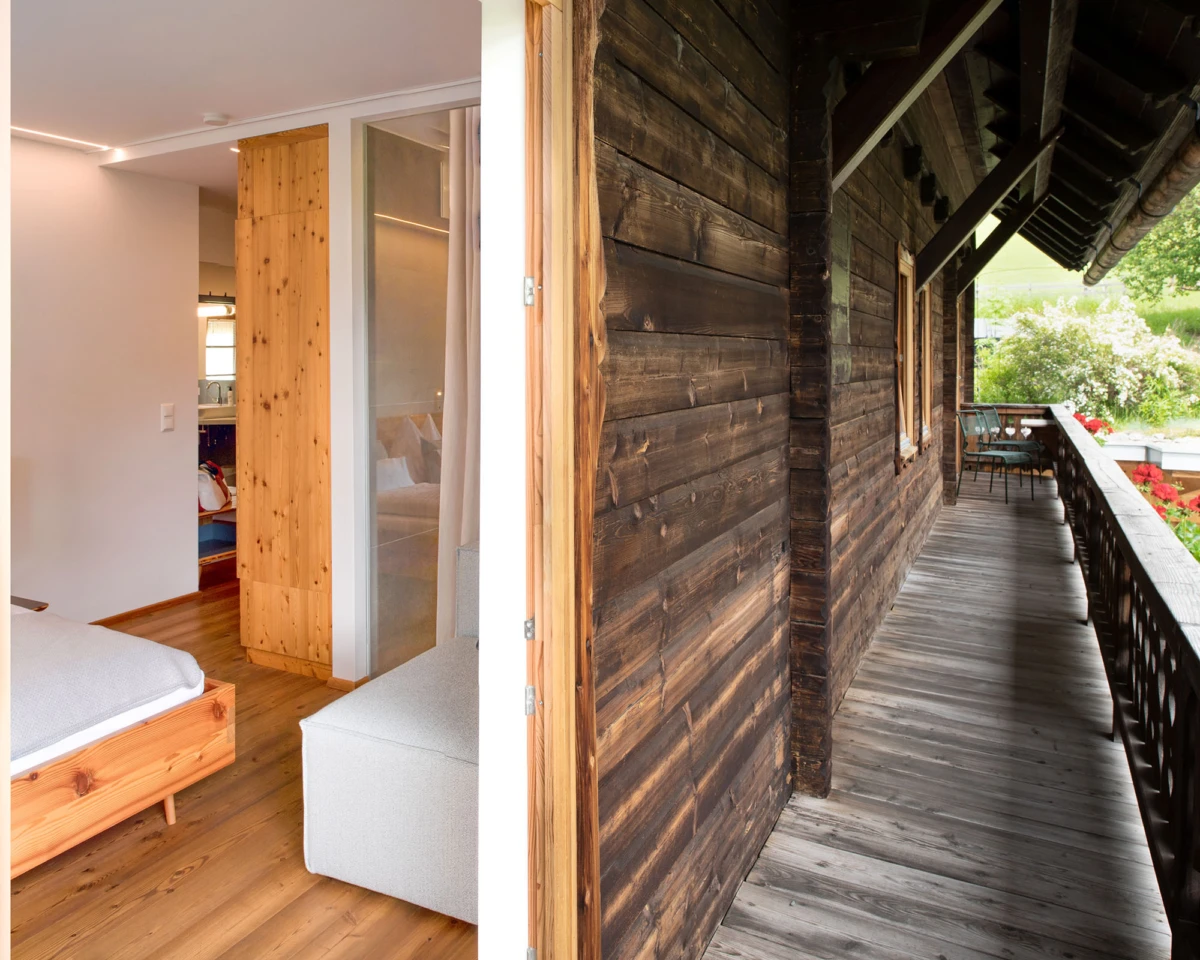 Innen- und Außenansicht eines Holzhauses: Schlafzimmer mit Bettgestell, Kleiderschrank und Sofa; Balkon mit Stühlen, Garten im Hintergrund.