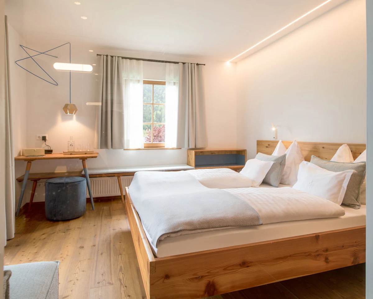 Massives Holzbett mit weißer Bettwäsche im hellen, modern eingerichteten Hotelzimmer; Arbeitsbereich mit Schreibtisch, Fenster mit Vorhängen.