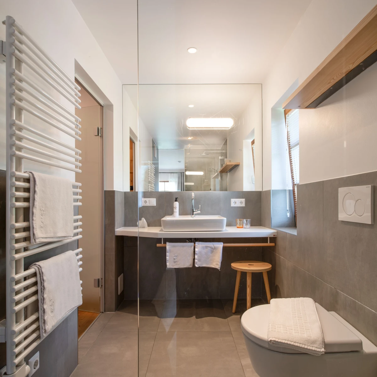 Modernes Badezimmer mit wandmontiertem Waschbecken, große Spiegelwand, Glasabtrennung zur Duschzone, Toilette, Holzhocker und weißem Handtuch.