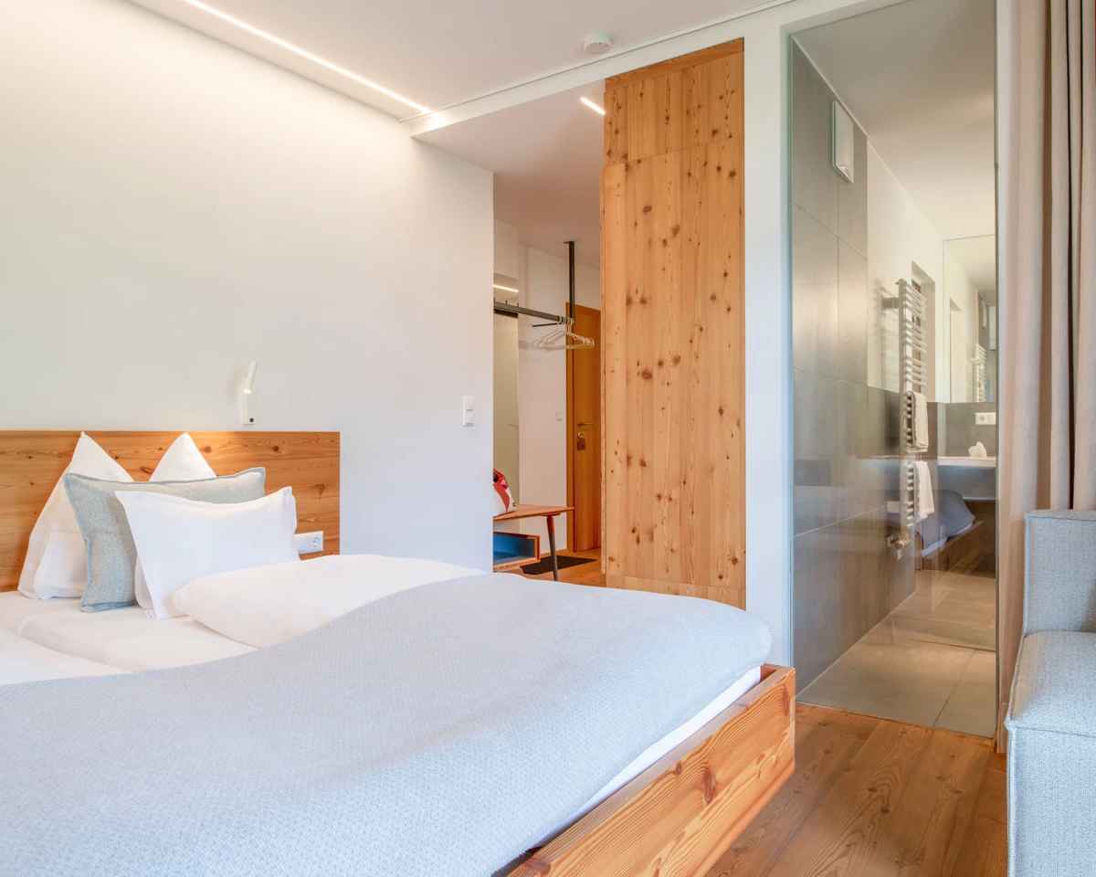 Hotelzimmer mit Doppelbett, graue Bettwäsche, Holzfußboden; angrenzendes Badezimmer mit Glaswand.