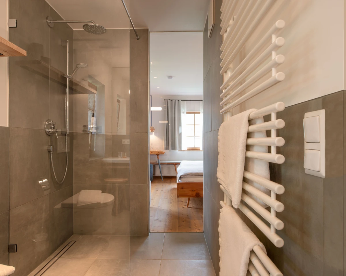 Modernes Badezimmer mit Glasdusche, Regendusche und Handbrause; rechter Heizkörper mit Handtüchern; Blick ins Schlafzimmer mit Holzboden und Bett.