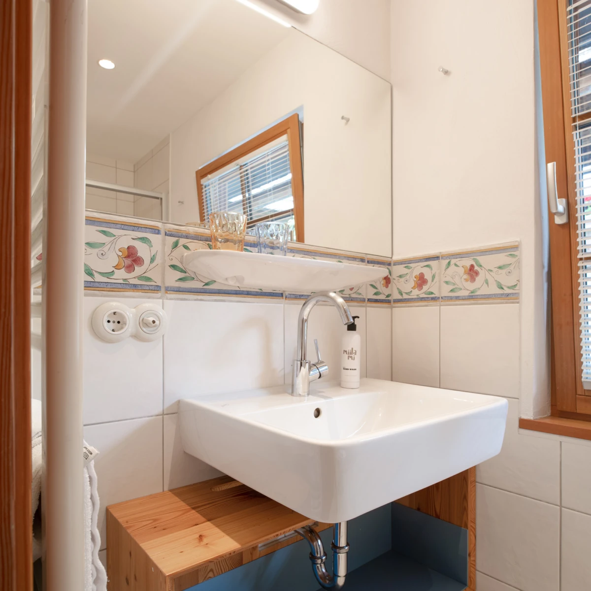 Waschbecken mit Armatur und Seifenspender vor blumiger Fliesenbordüre; Spiegel darüber, Fenster rechts.