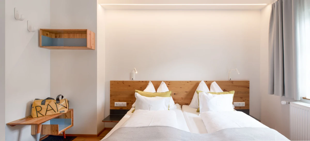 Hotelzimmer mit zwei Einzelbetten, weißer Bettwäsche und grünen Kissen, Holzkopfteil, Nachttische, Wandlampen, Fenster mit grauen Vorhängen.