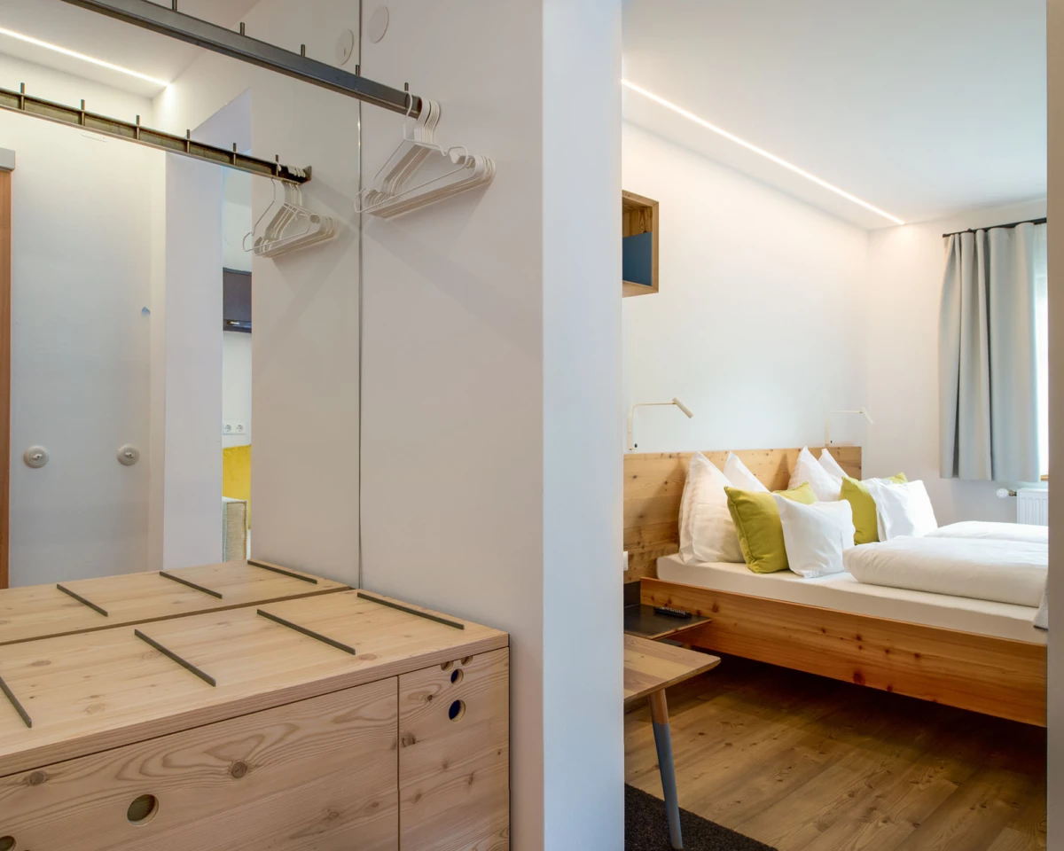 Schlafzimmer mit massivem Holzbett, Bettwäsche in Weiß und grünen Akzentkissen; linker Kleiderschrankbereich.