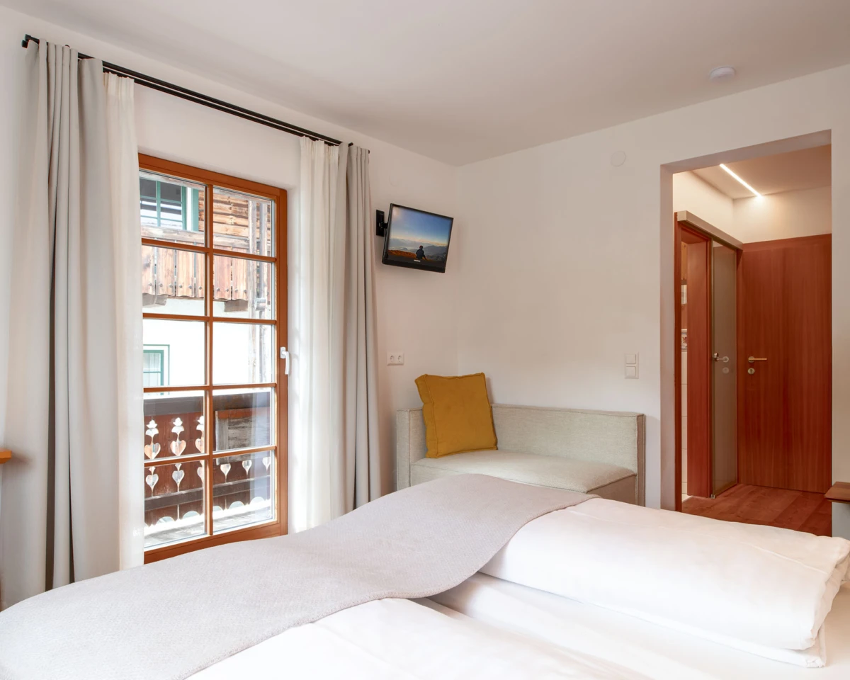 Hotelzimmer mit Doppelbett, Fenster mit Vorhängen, Wandfernseher, Sofa mit senfgelbem Kissen, offener Durchgang zum Flur.