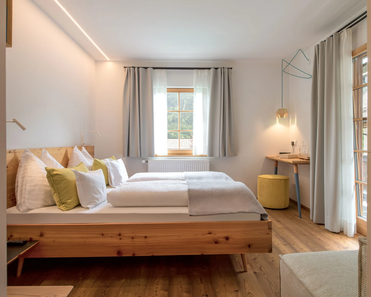 Doppelbett aus Massivholz mit weißer Bettwäsche und grünen Kissen; Arbeitsbereich und großem Fenster mit grauen Vorhängen.
