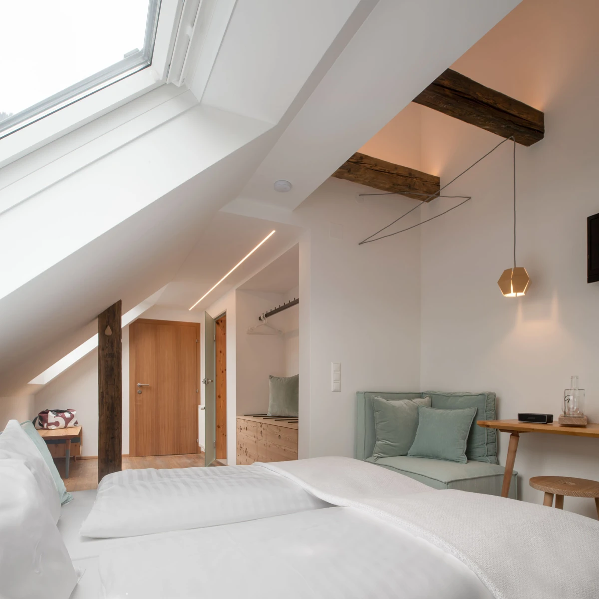 Schlafzimmer im Dachgeschoss mit Dachfenster, sichtbaren Holzbalken, weißem Bett und Sitzecke mit mintgrünen Kissen.