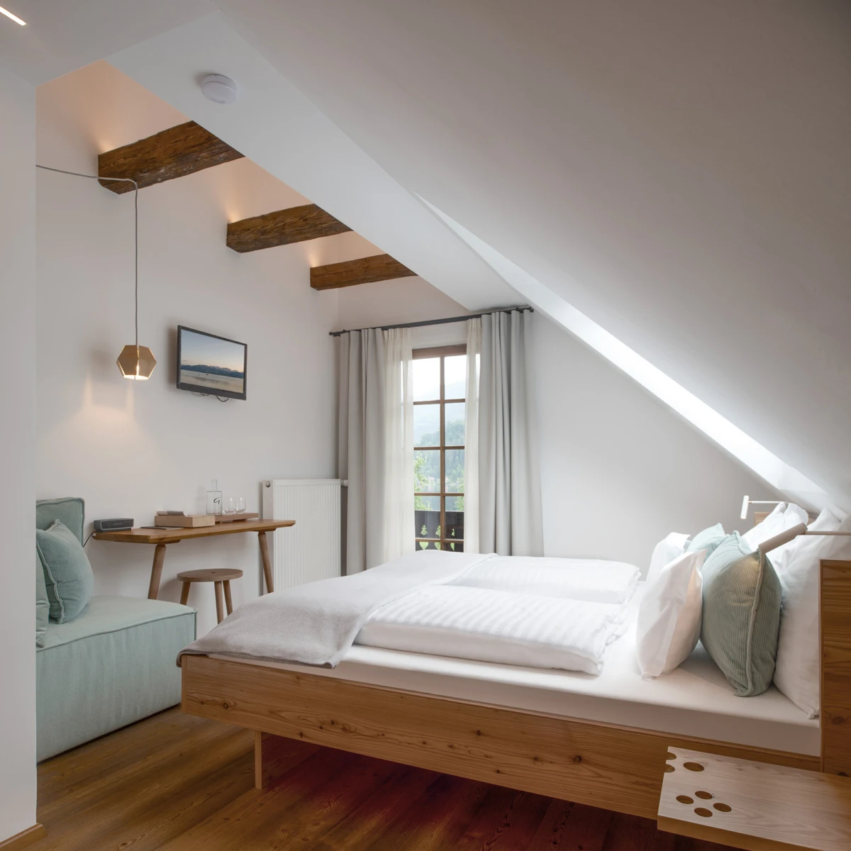 Schlafzimmer mit schräger Decke und Holzbalken, Doppelbett, weiße Bettwäsche, Sofa, Schreibtisch, Wandfernseher, Fenster mit Vorhängen.