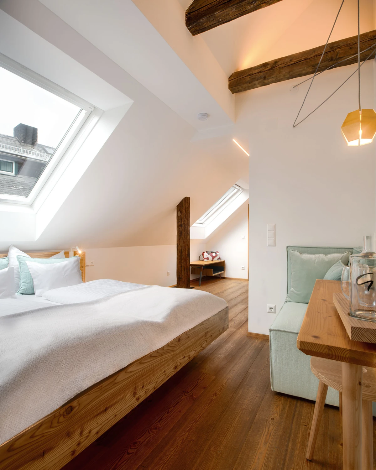 Doppelbett im hellen Dachgeschosszimmer mit zwei Dachfenstern, sichtbaren Holzbalken, Holzfußboden und Sitzecke mit Holztisch.