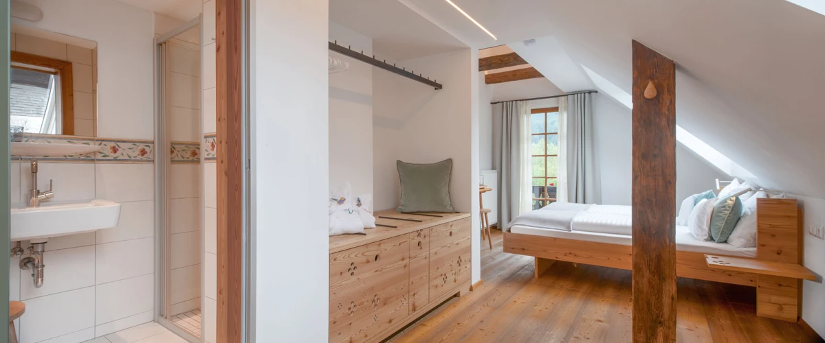 Loftartiges Attikazimmer mit Doppelbett, Holzboden, sichtbaren Holzbalken, Kommode und Badezimmer links mit Waschbecken.