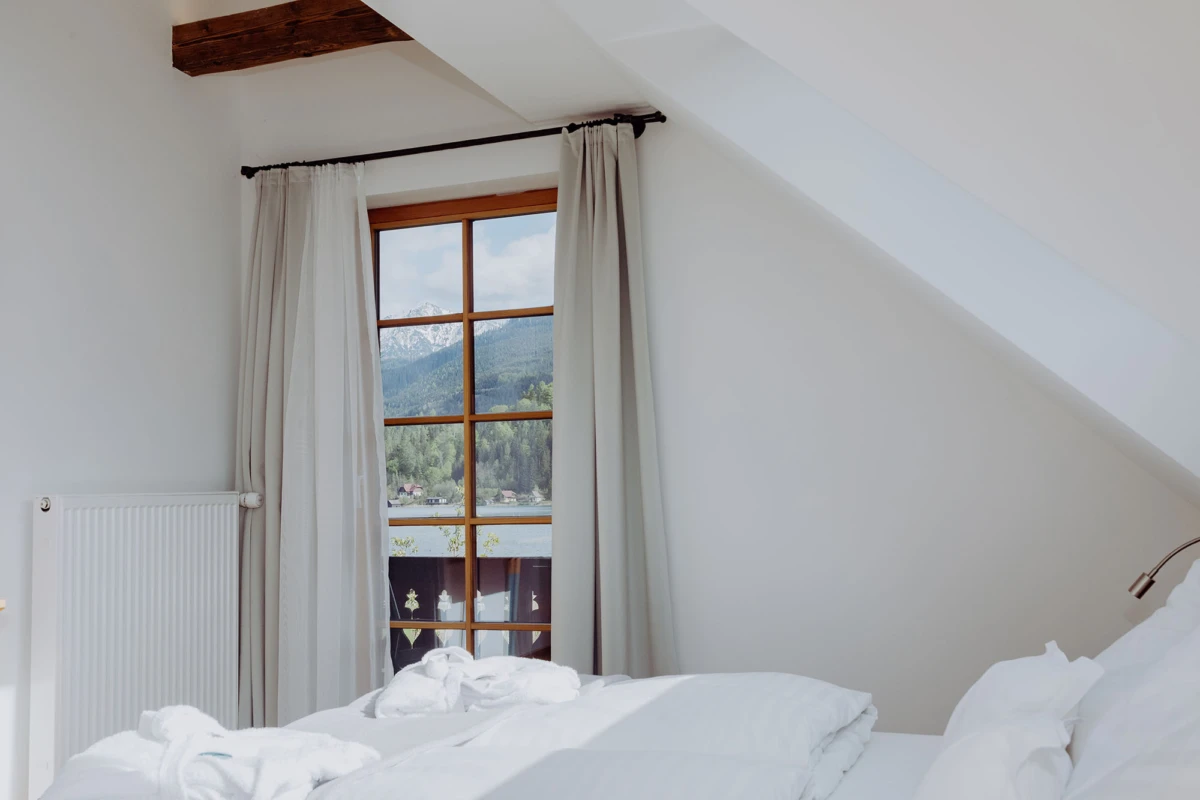 Schlafzimmer mit weißem Bett, schräger Decke; Holzrahmenfenster mit Vorhängen, Blick auf Bergwald und Berge.