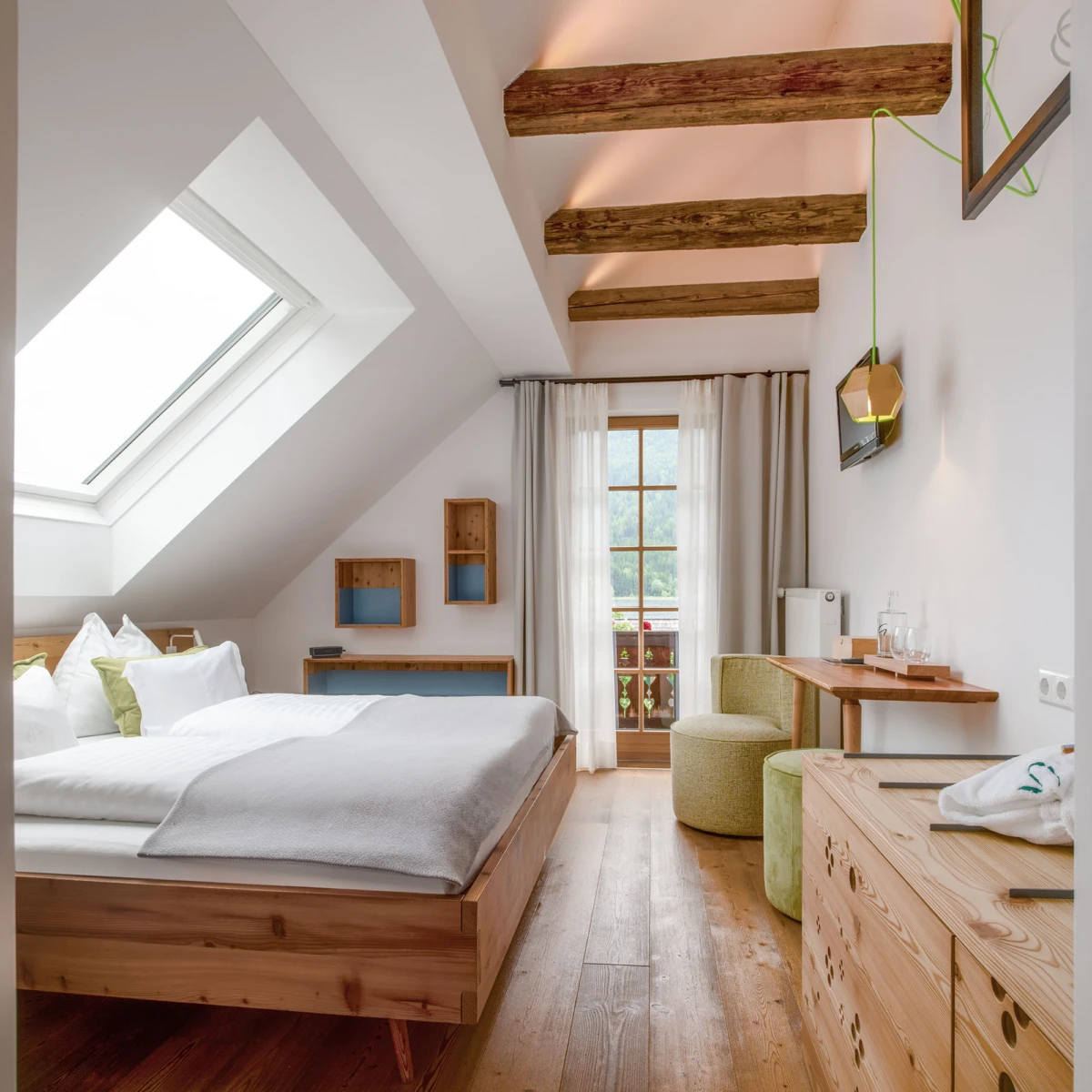 Massivholz-Doppelbett im hellen Schlafzimmer mit Decke aus Holzbalken, Dachfenster, Balkonzugang, hellen Vorhängen und Holzfußboden.