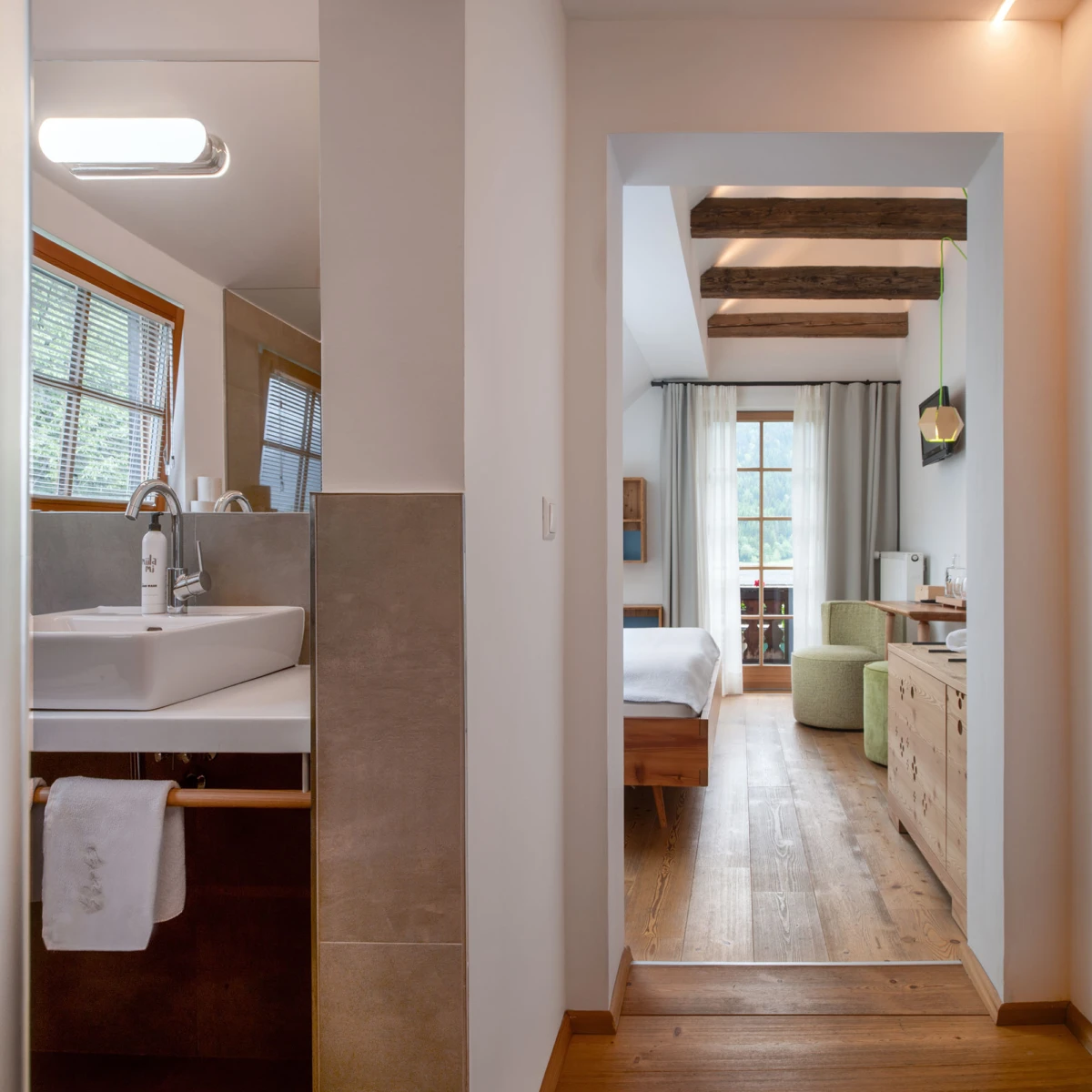 Helles Hotelzimmer-Suite-Durchgang: Badezimmer mit Waschbecken links, Schlafzimmer mit Bett und Fenster rechts, Holzbalkendecke, Holzboden.